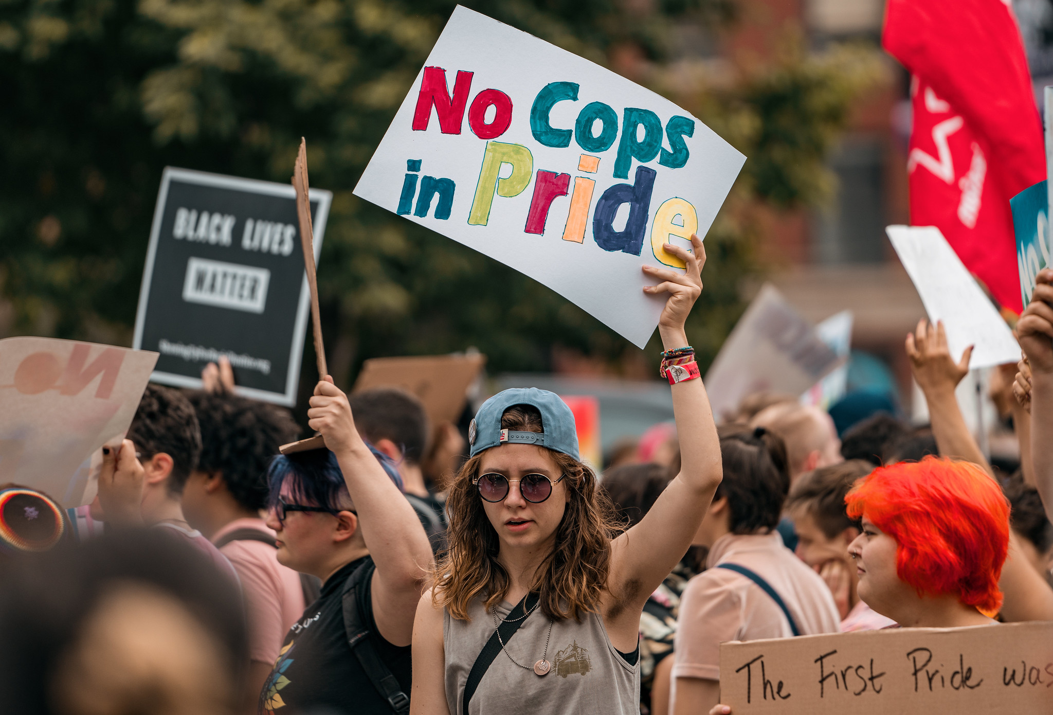 a person in a gray shirt and backwards baseball hat holds a colorful handwritten sign that says "no cops in pride"