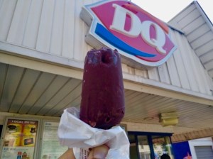A hand holds a chocolate-coated Dilly Bar next to the DQ logo