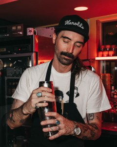 portrait of bennett johnson, who's mixing a drink and wearing a "Harold's" baseball cap