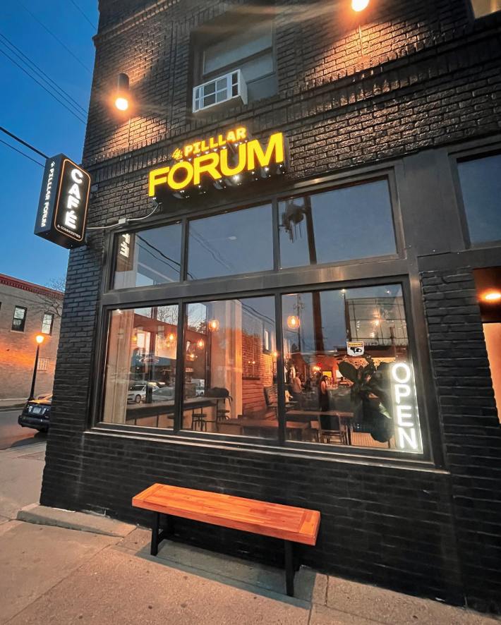 A nighttime view of Pilllar Forum Cafe and Commodities, with the sign lit up and people sitting inside