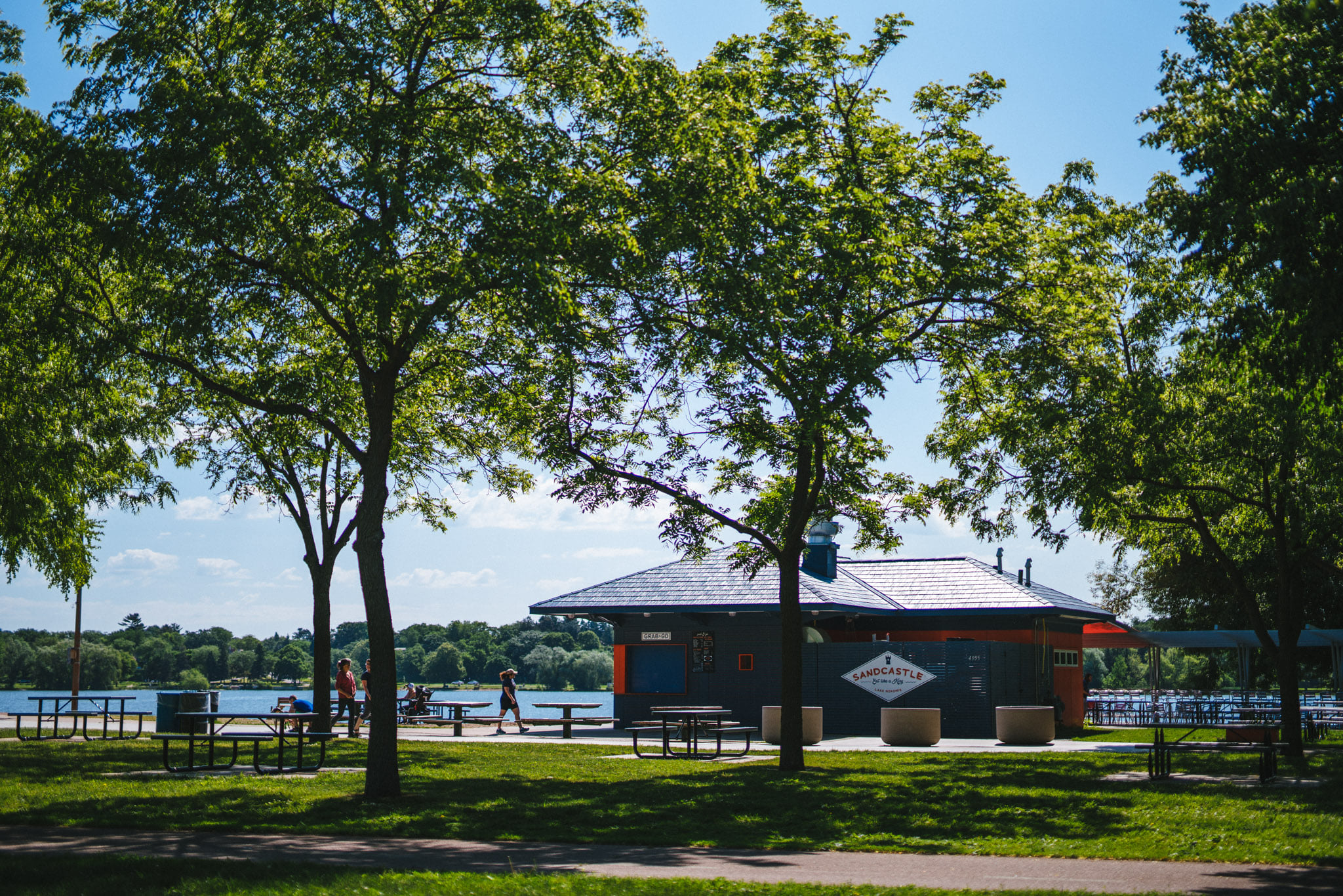 An exterior shot of Sandcastle, with lush green trees in the foreground and Lake Nokomis in the background