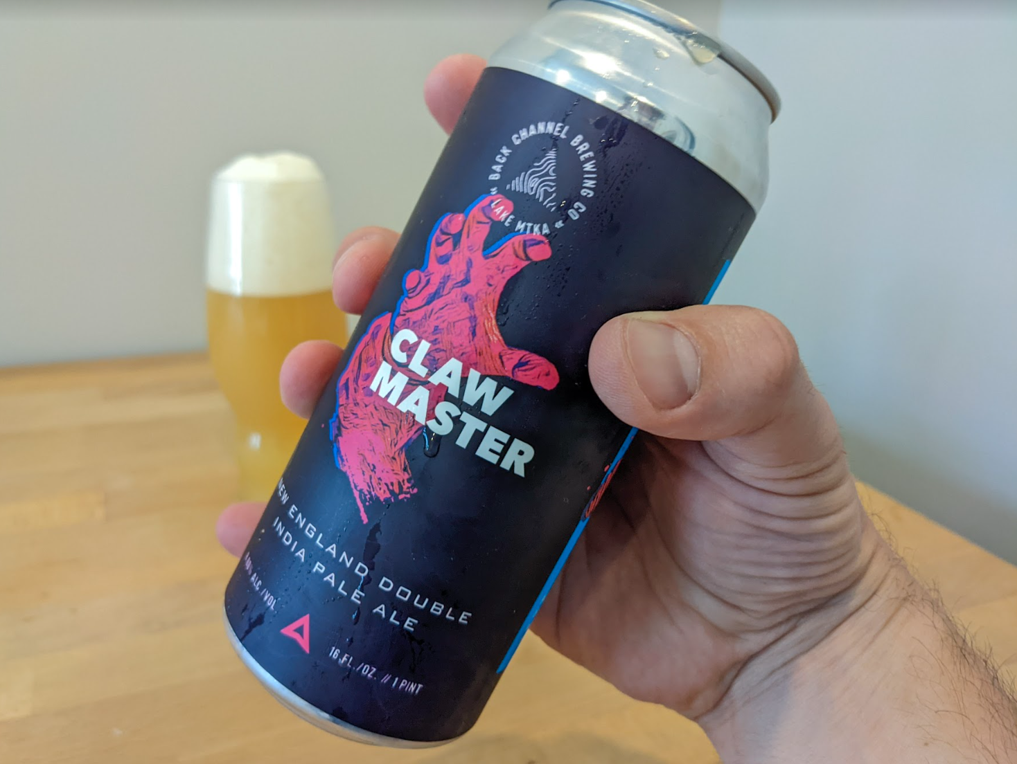 a hand holds claw master's black and red can, while a beer sits on a table in the background