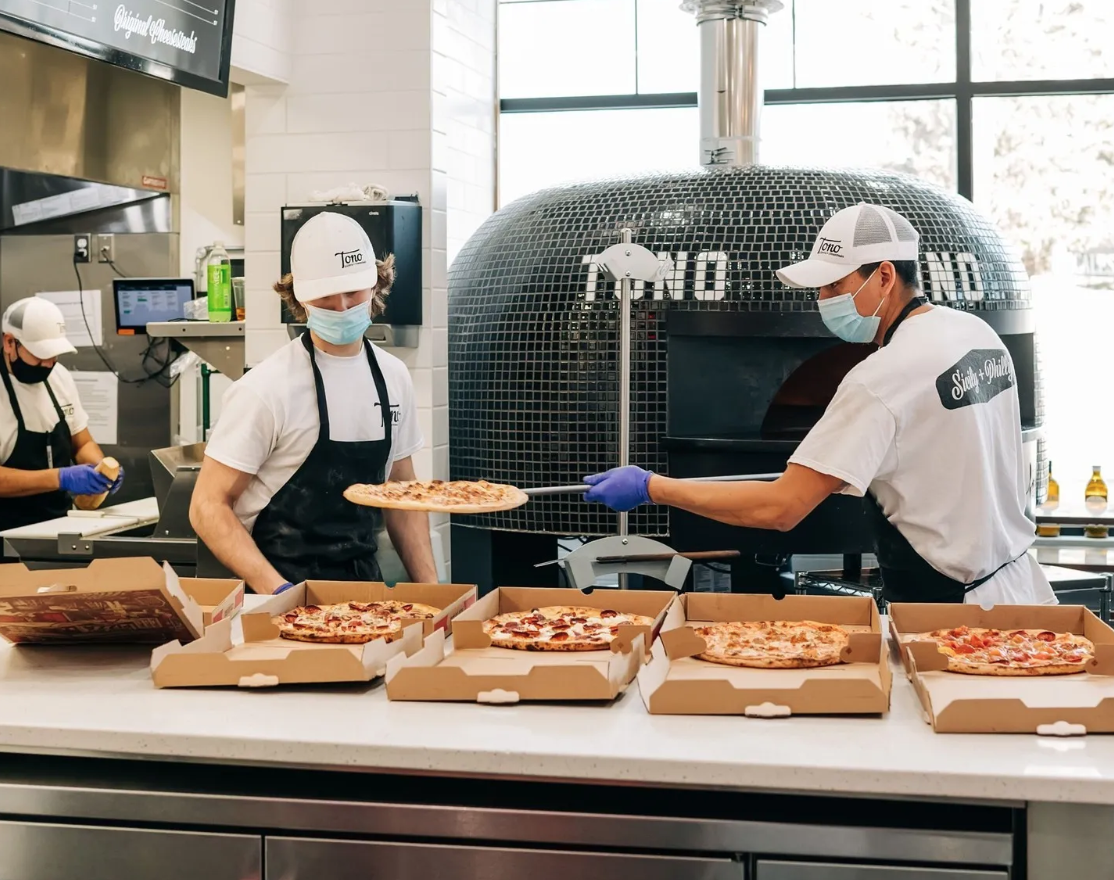 two men in front of a big tono oven slide pizzas off of paddles into boxes in front of them