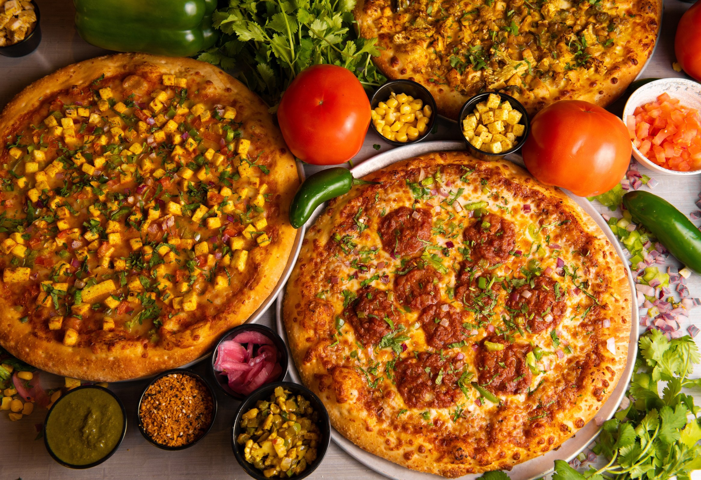 an overhead shot of three colorful Bombay Pizza Kitchen pies, surrounded by ingredients
