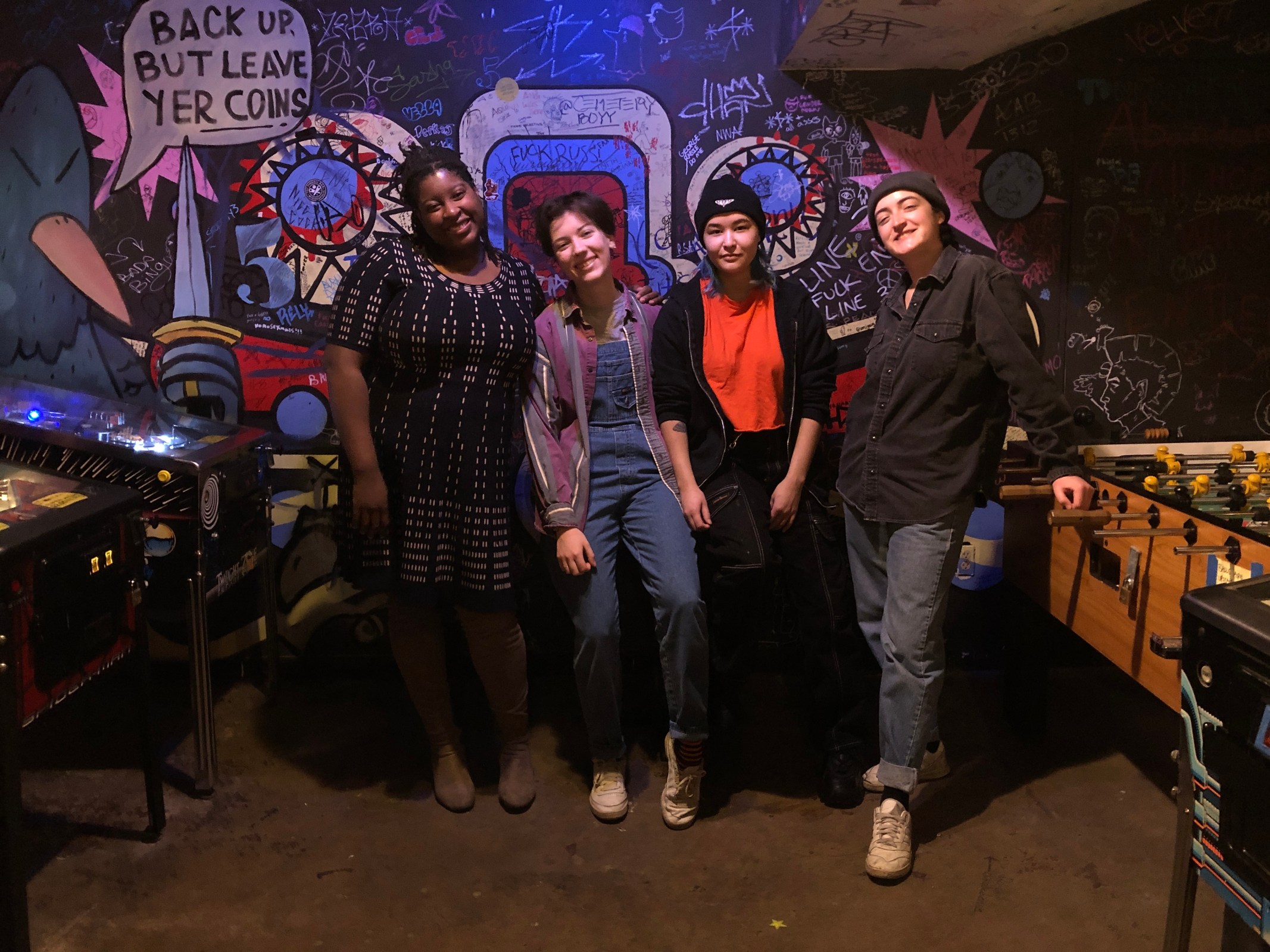 four people stand smiling next to the pinball machines in the basement of Cafetto