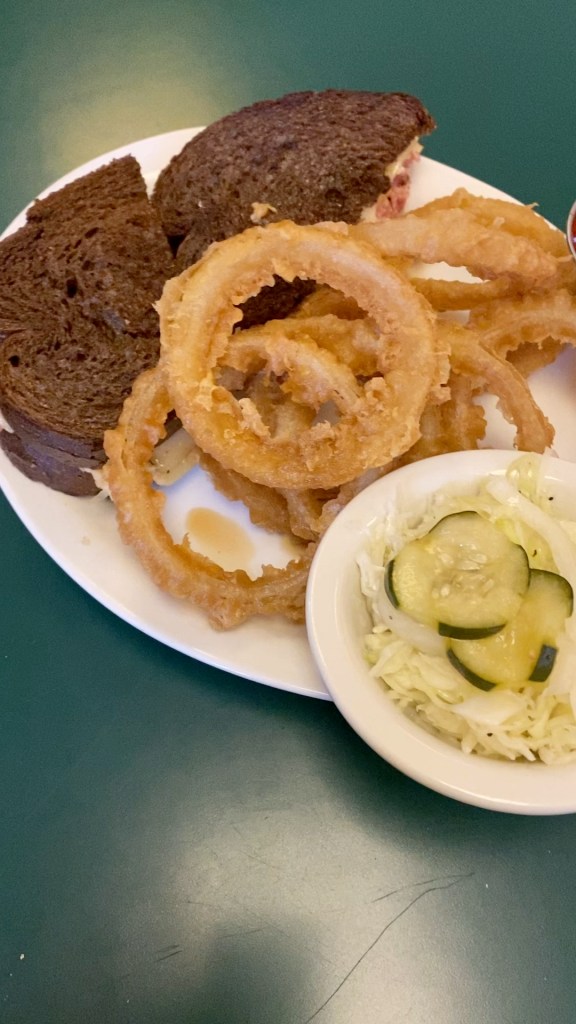 a reuben sandwich on rye, next to a plate of onion rings and pickles