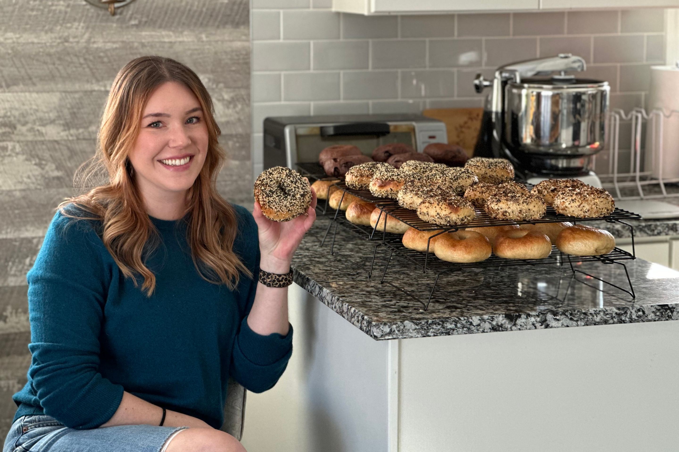 megan of mogi bagel holds an everything bagel while seated next to rows and rows of other bagels