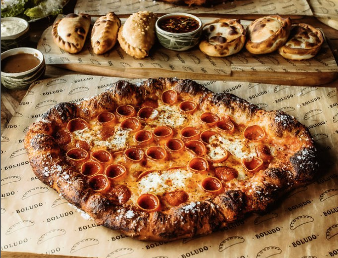 foreground: one of boludo's beautiful diamond-shaped pies, with aa charred, salt-dusted crust and cupped pepperonis throughout. background: a plank with six toasty empanadas, with dipping sauce in the center.