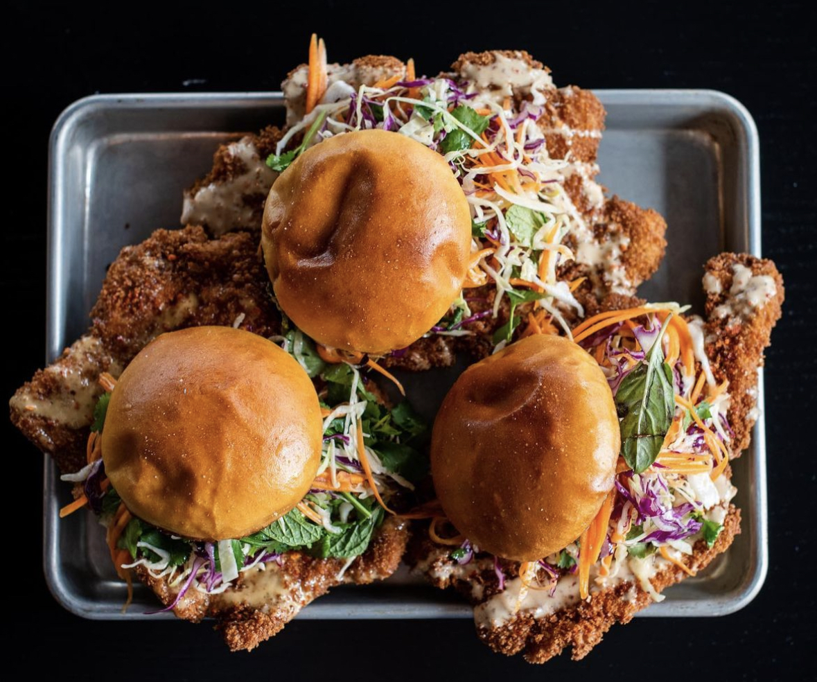 An overhead shot of three enormous pork tenderloin sandwiches, which are topped with colorful slaw and wear little buns like hats