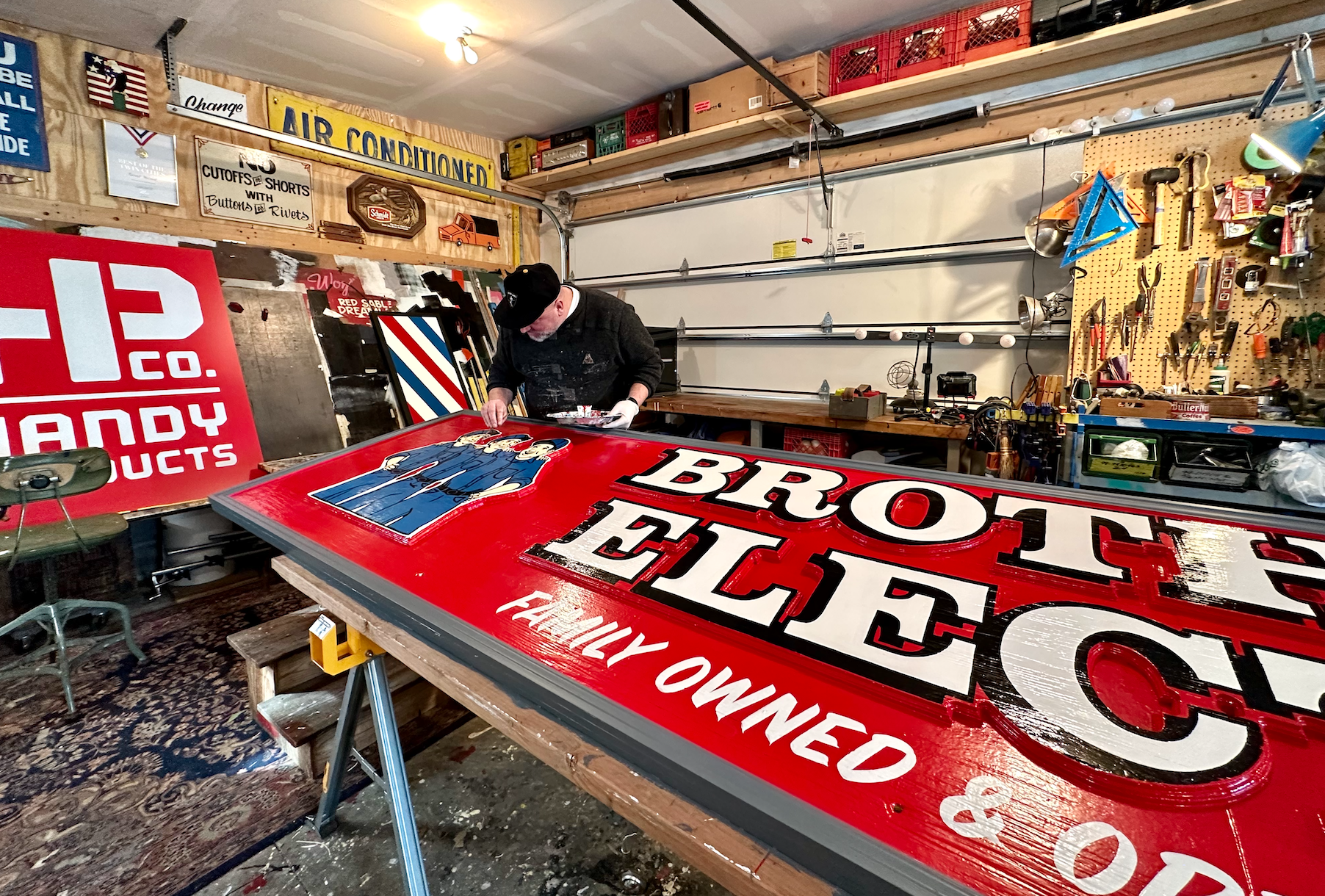 a man paints a bright red sign reading "brothers electric" in a garage studio space
