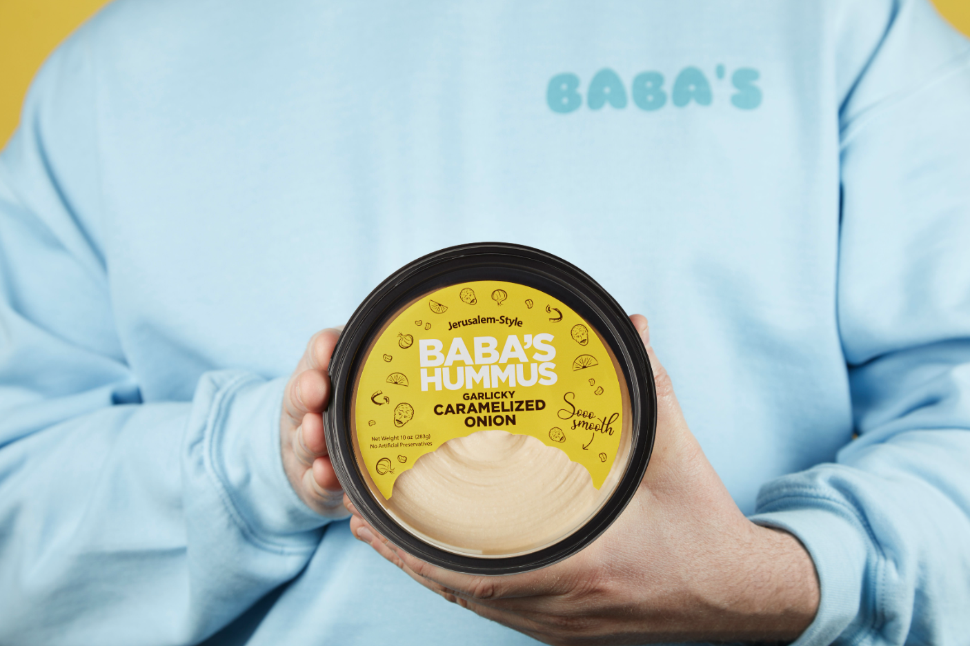 a person in a baby blue baba's sweatshirt holds their garlicky onion hummus to the camera