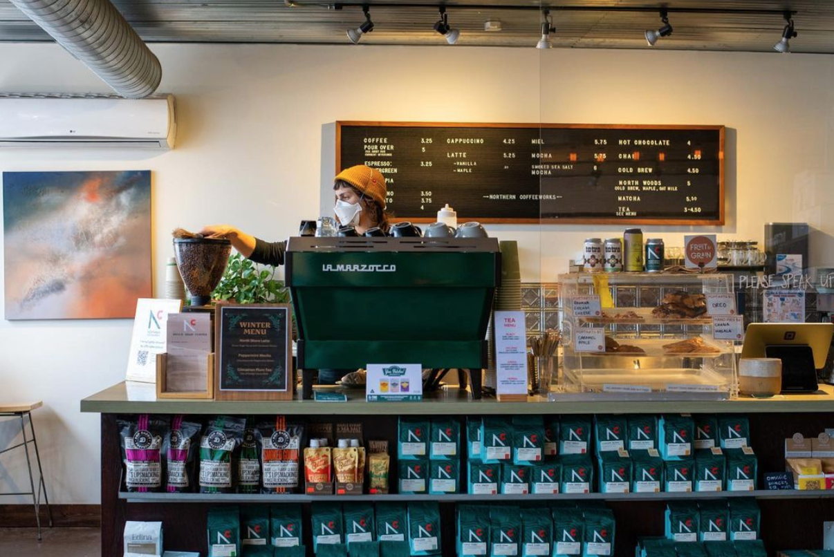 A barista in a yellow beanie prepares coffee behind a counter stocked with backs of beans and coffee equipment
