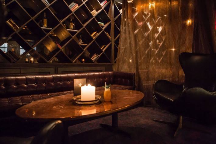 A dark wooden table surrounded by deep maroon and black leather seating, with semi-sheer curtains separating the area from the rest of the bar