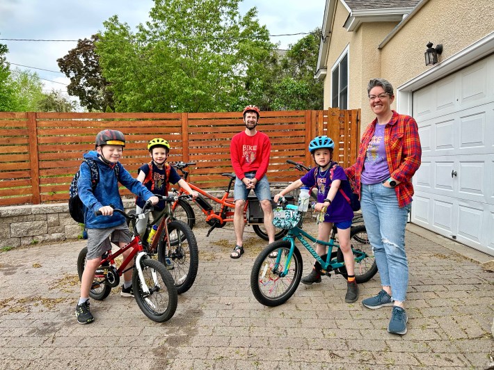 Two adults and three children, all in colorful bike bus shirts designed by a local illustrator, wait in their driveway for the go-ahead to start riding