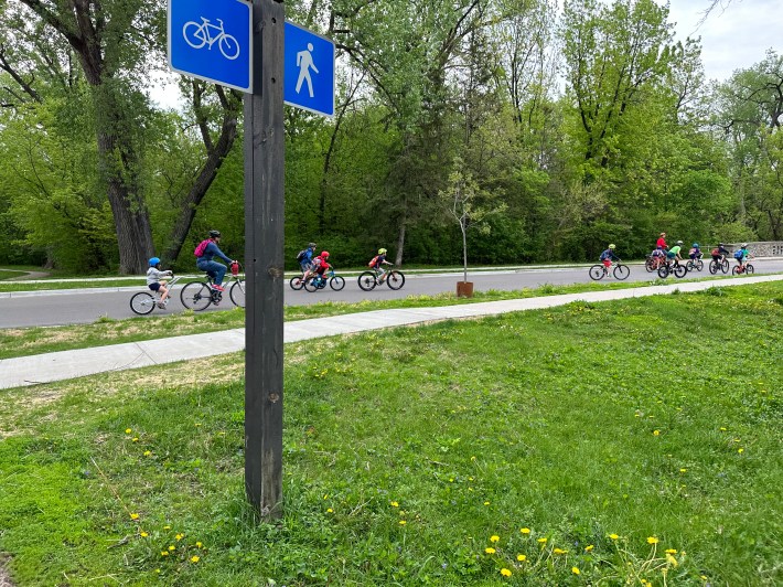 in the foreground, a blue shared roadway sign with a graphic of a bike on one side and a pedestrian on the other. in the background, a group of children rides their bikes up a hill.