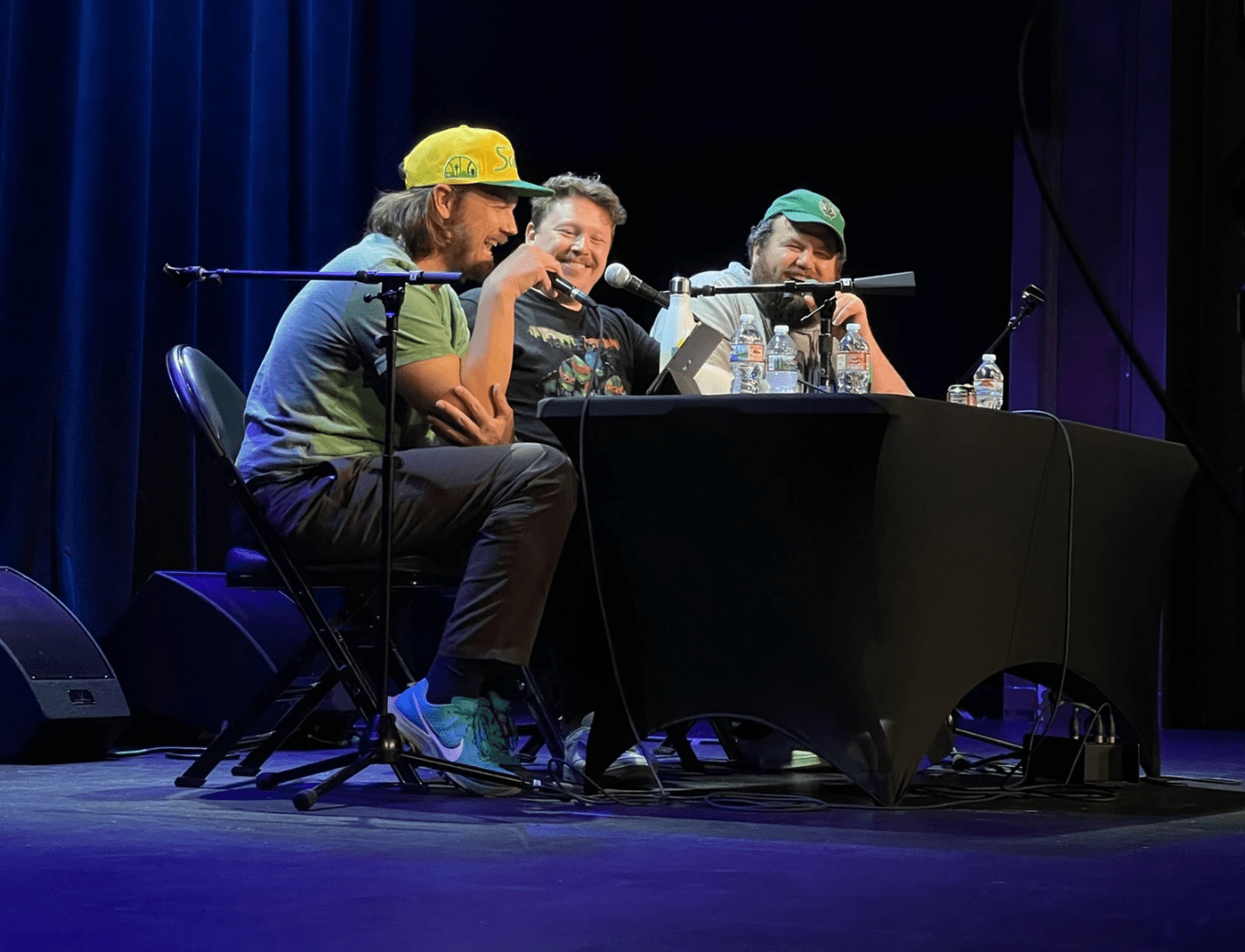 three men sit at a table on a stage laughing with one another
