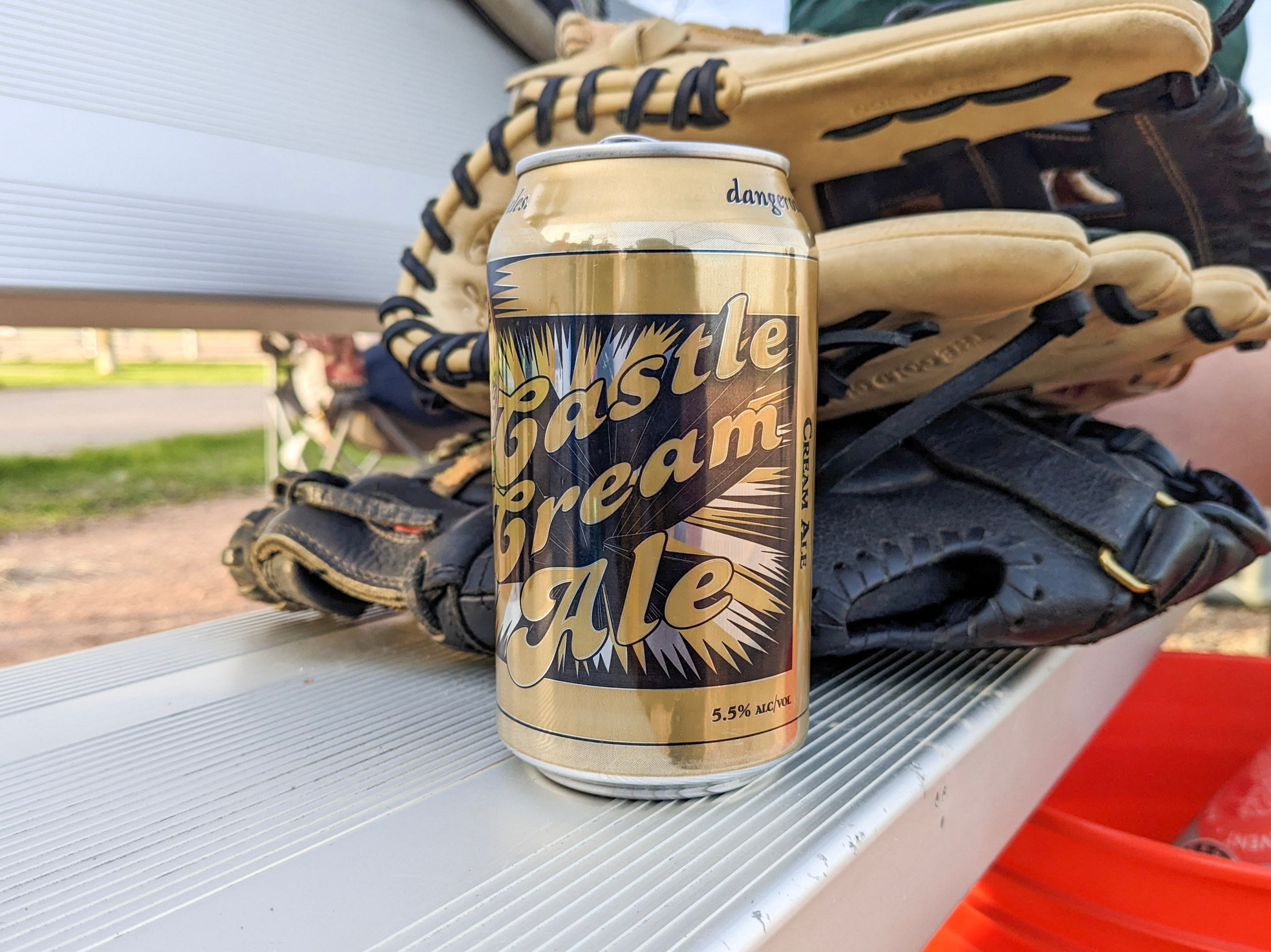 A golden can of cream ale sits on a metal bleacher in front of some softball gloves